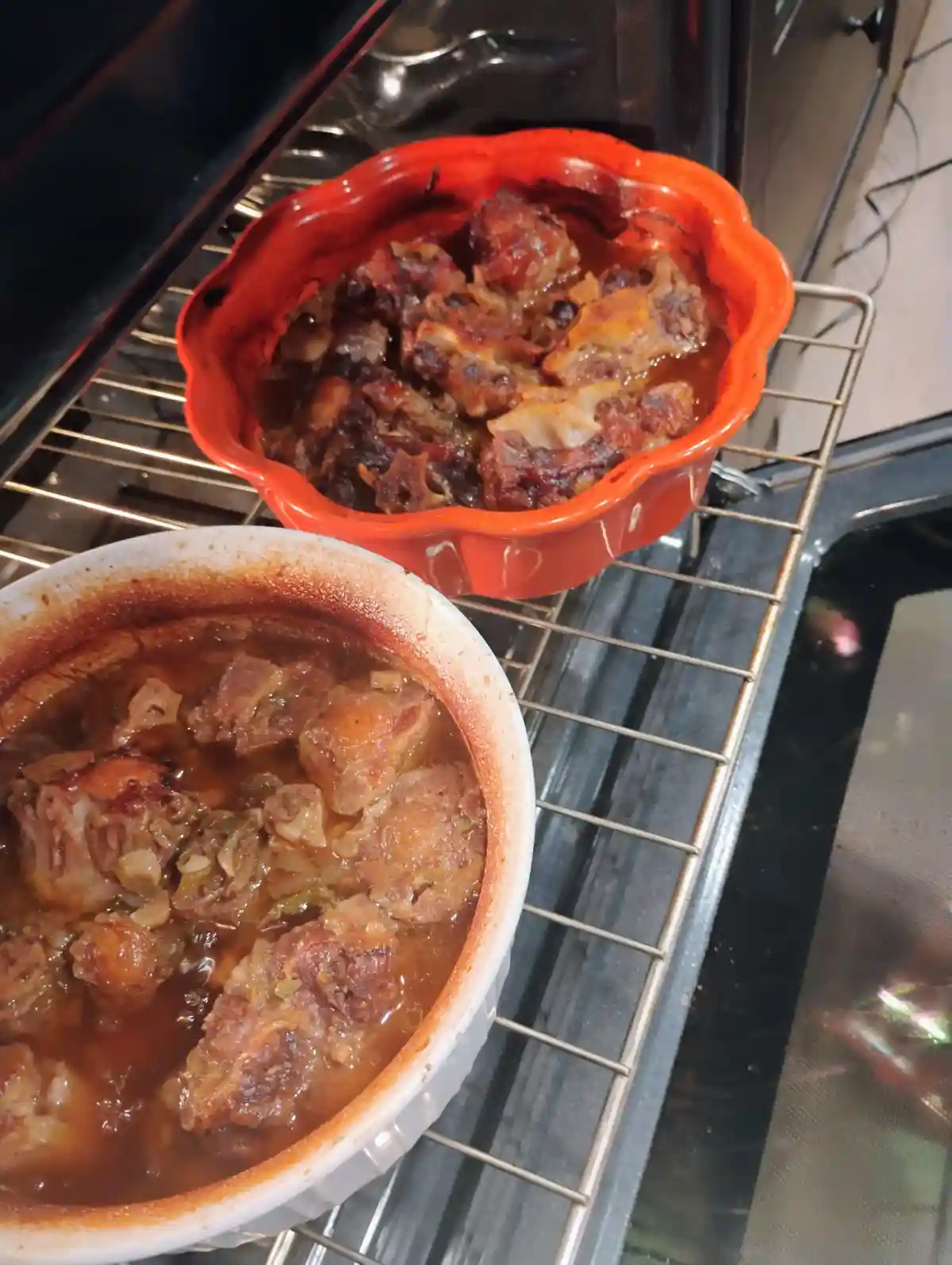 Two ceramic dishes filled with cooked meat and sauce sit on an oven rack; one dish is orange and appears burnt, the other is…
