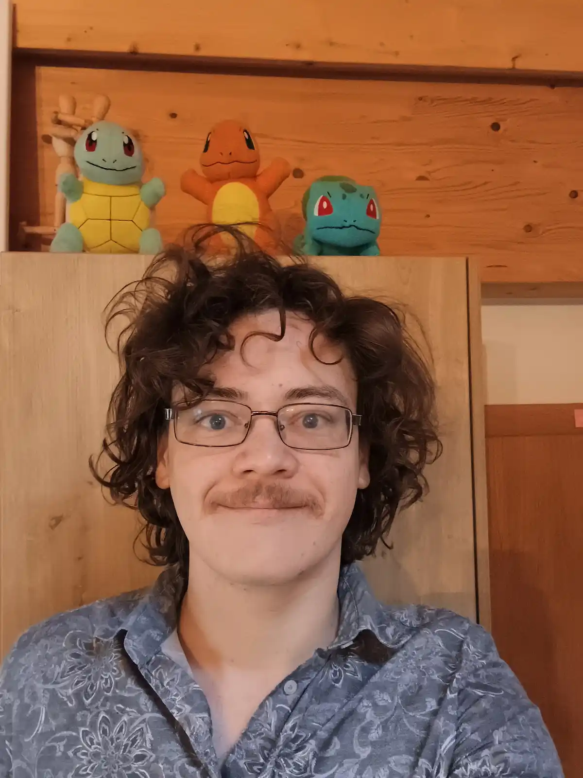 A person with curly brown hair and glasses smiles at the camera while wearing a blue patterned shirt, with three Pokémon…