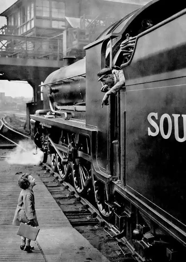 A vintage black-and-white photo shows a young child standing on a train platform holding a suitcase, looking up at a steam…