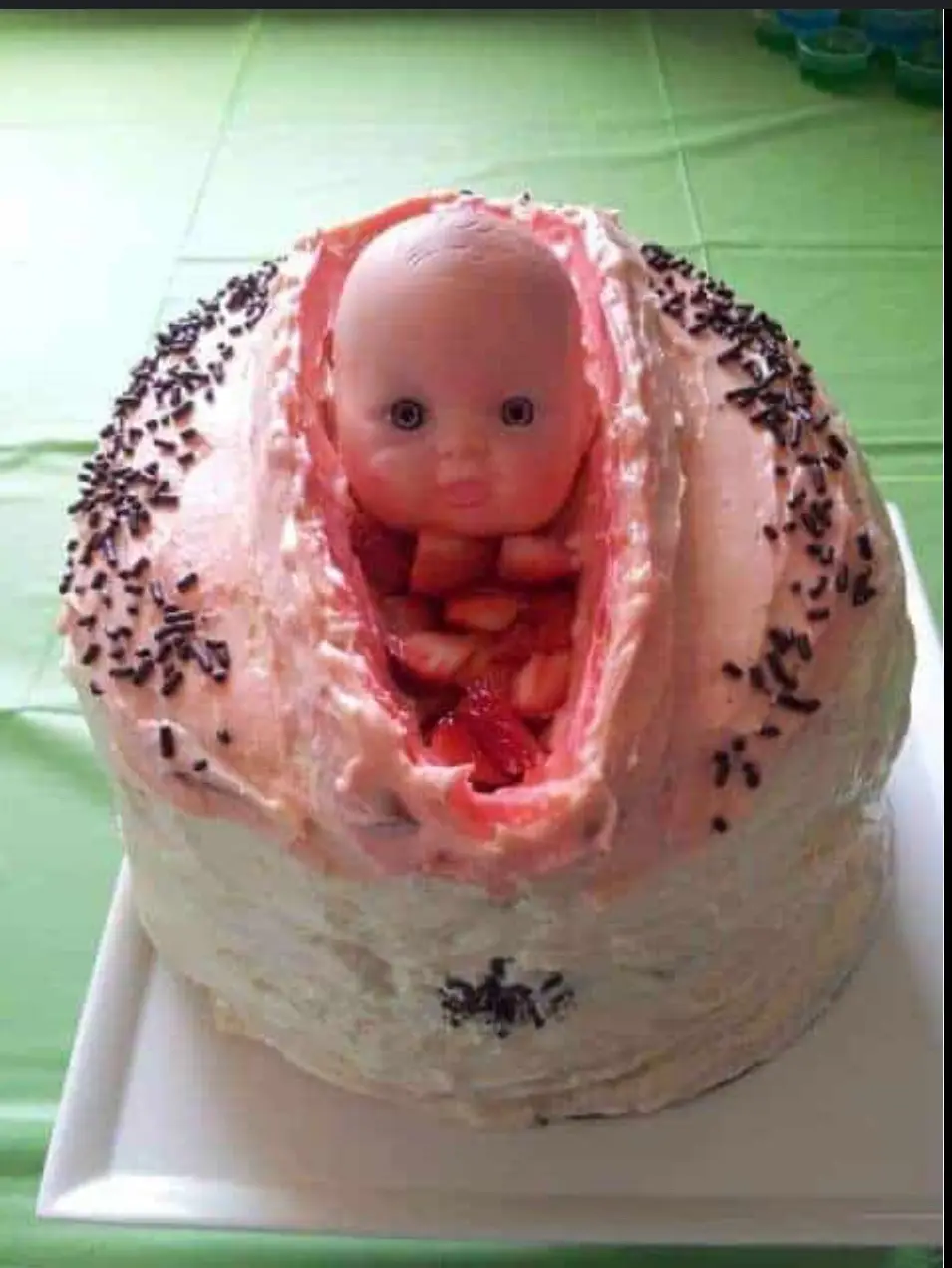 A round cake decorated with pink and white frosting and chocolate sprinkles, designed to look like a baby emerging from a…