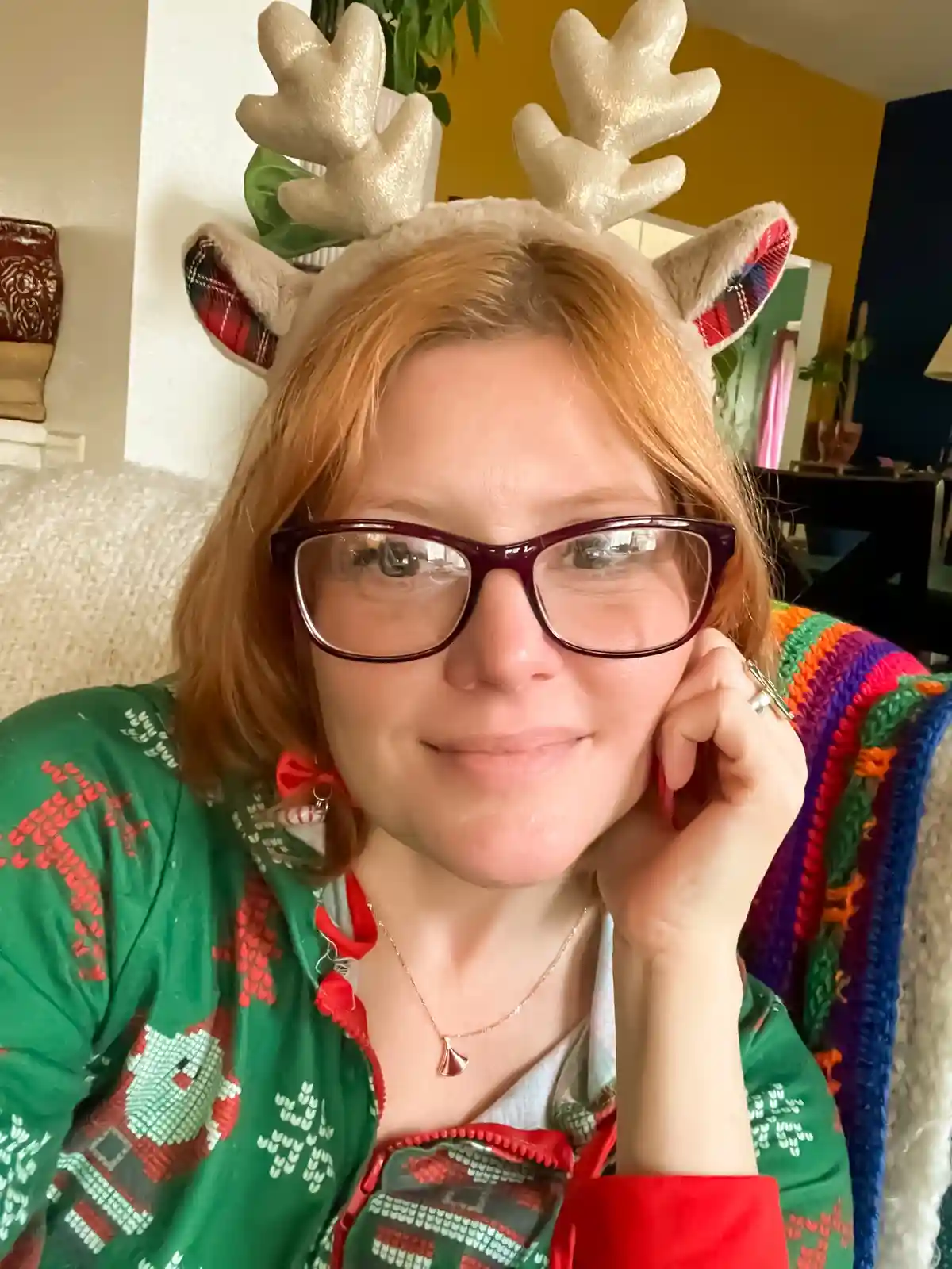 A person with reddish hair and glasses wears a sparkly reindeer antler headband with plaid ears, smiling while resting their…
