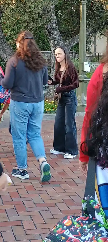 A woman in a striped shirt and jeans stands outdoors on a brick walkway, looking visibly annoyed or disappointed at someone…