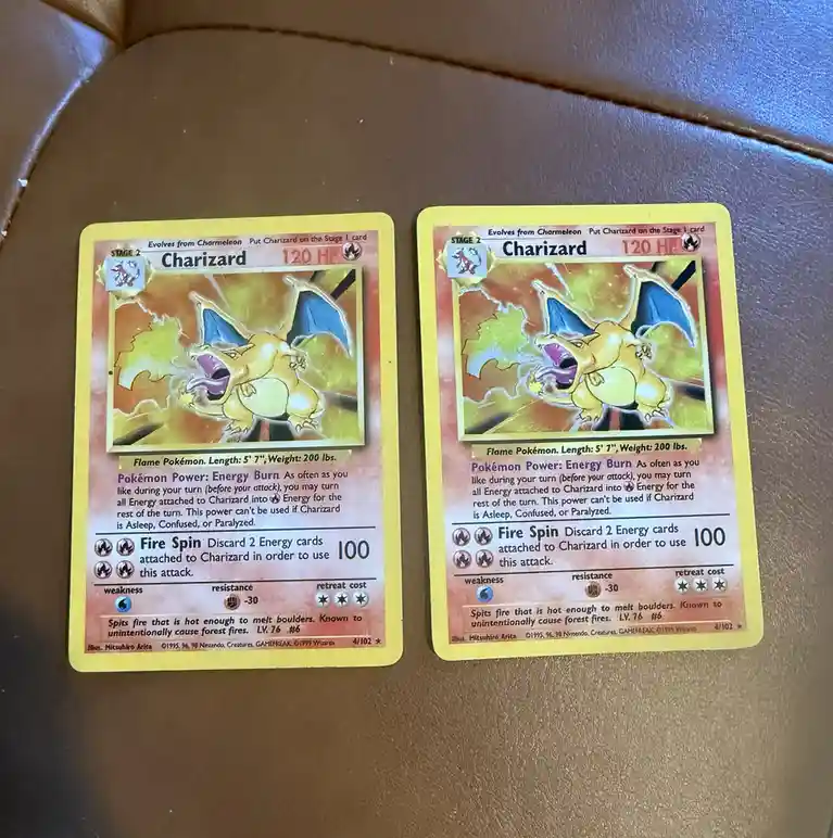 Two identical Pokémon Trading Card Game Charizard cards are placed side-by-side on a brown leather surface, showing the same…