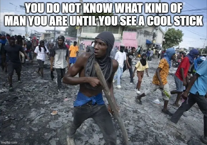 A shirtless Black man wearing a grey head covering and blue pants stands in the foreground of a chaotic street scene…