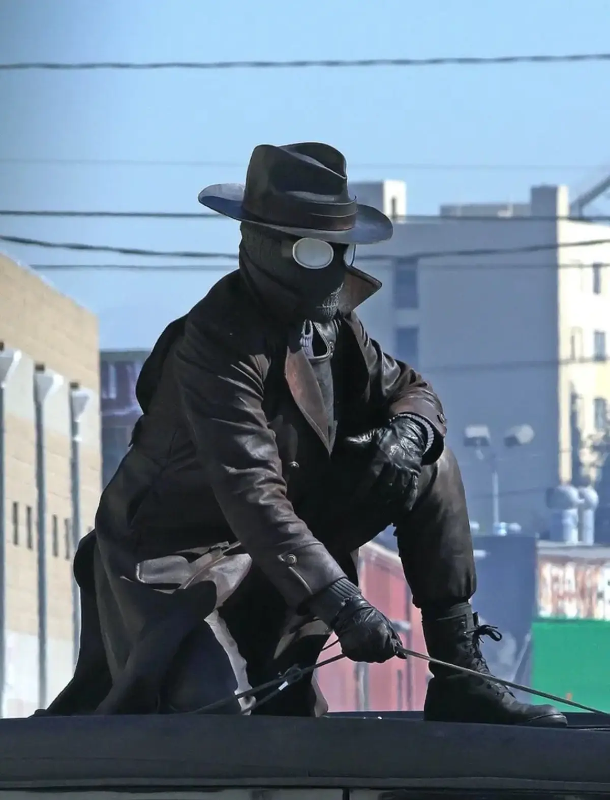 A person in a dark fedora, leather trench coat, and a skull mask with a single round eyepiece crouches on a rooftop, holding…