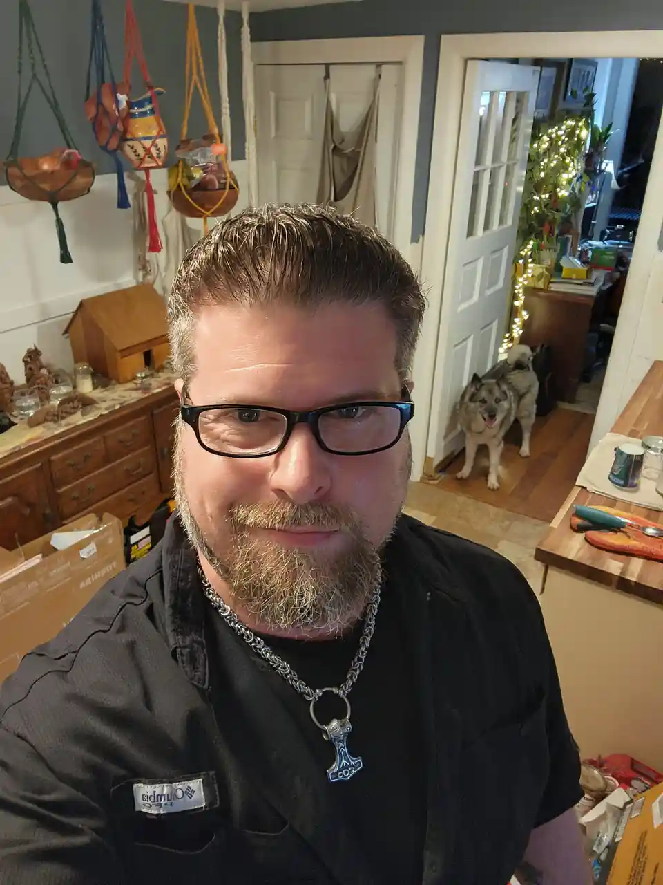 A man with glasses and a beard wears a silver Thor's hammer pendant on a chain, smiling at the camera in a home setting…