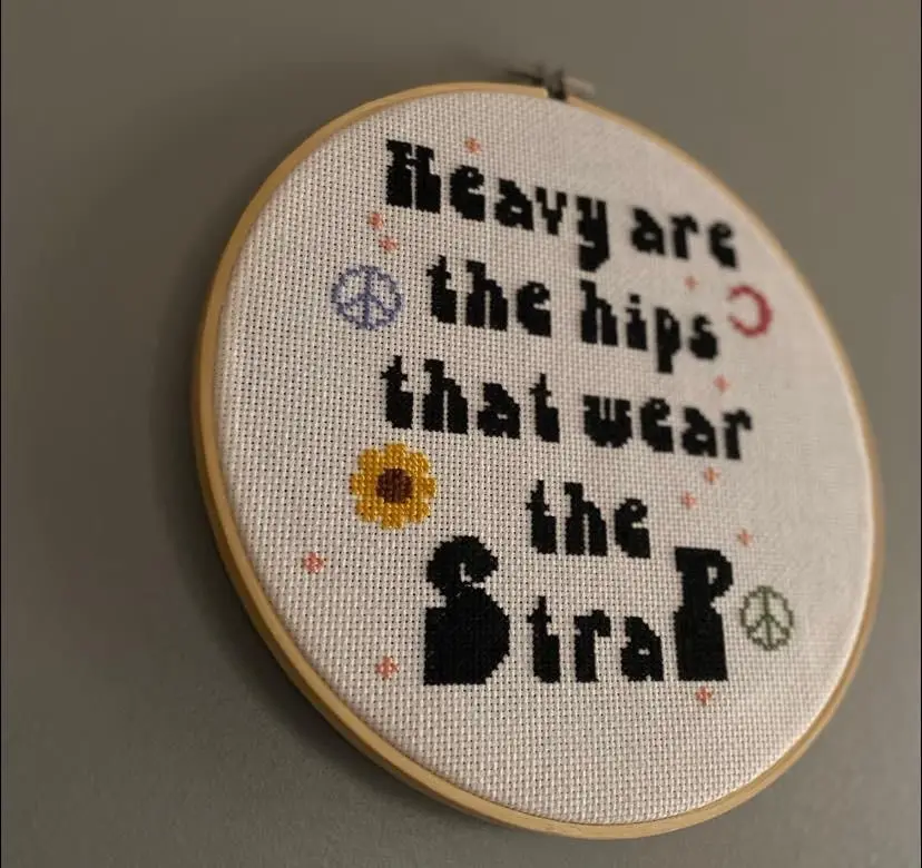 A circular embroidery hoop displays a cross-stitch pattern with the text 'Heavy are the hips that wear the strap' in black…