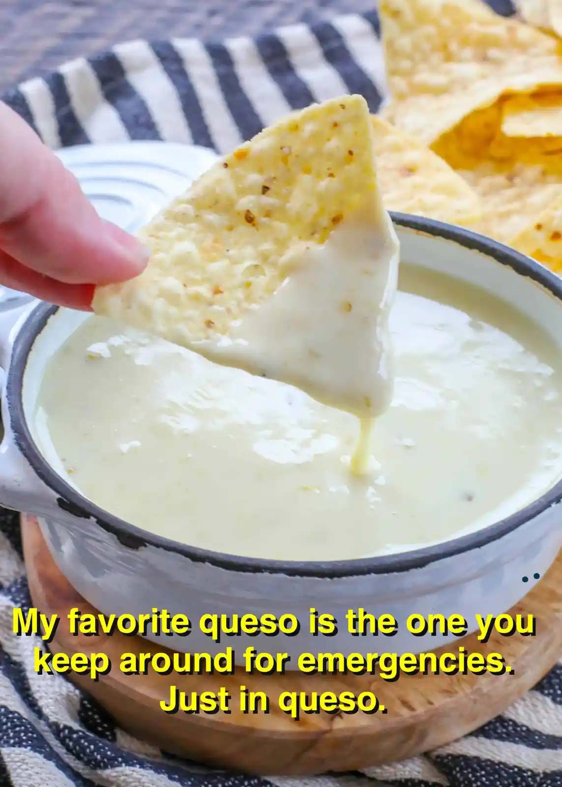 A hand dips a triangular tortilla chip into a bowl of creamy white queso dip, with more chips visible in the background. The…