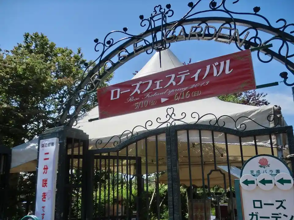 An ornate black metal gate with 'ROSE GARDEN' written across the top arch, under which hangs a red banner announcing…