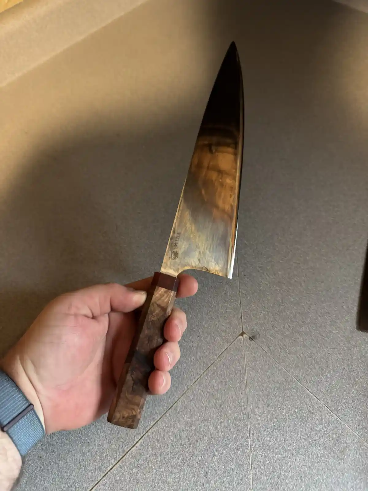 A hand holds a large chef's knife with a wooden handle and a worn, reflective blade over a gray tiled surface. The overlaid…