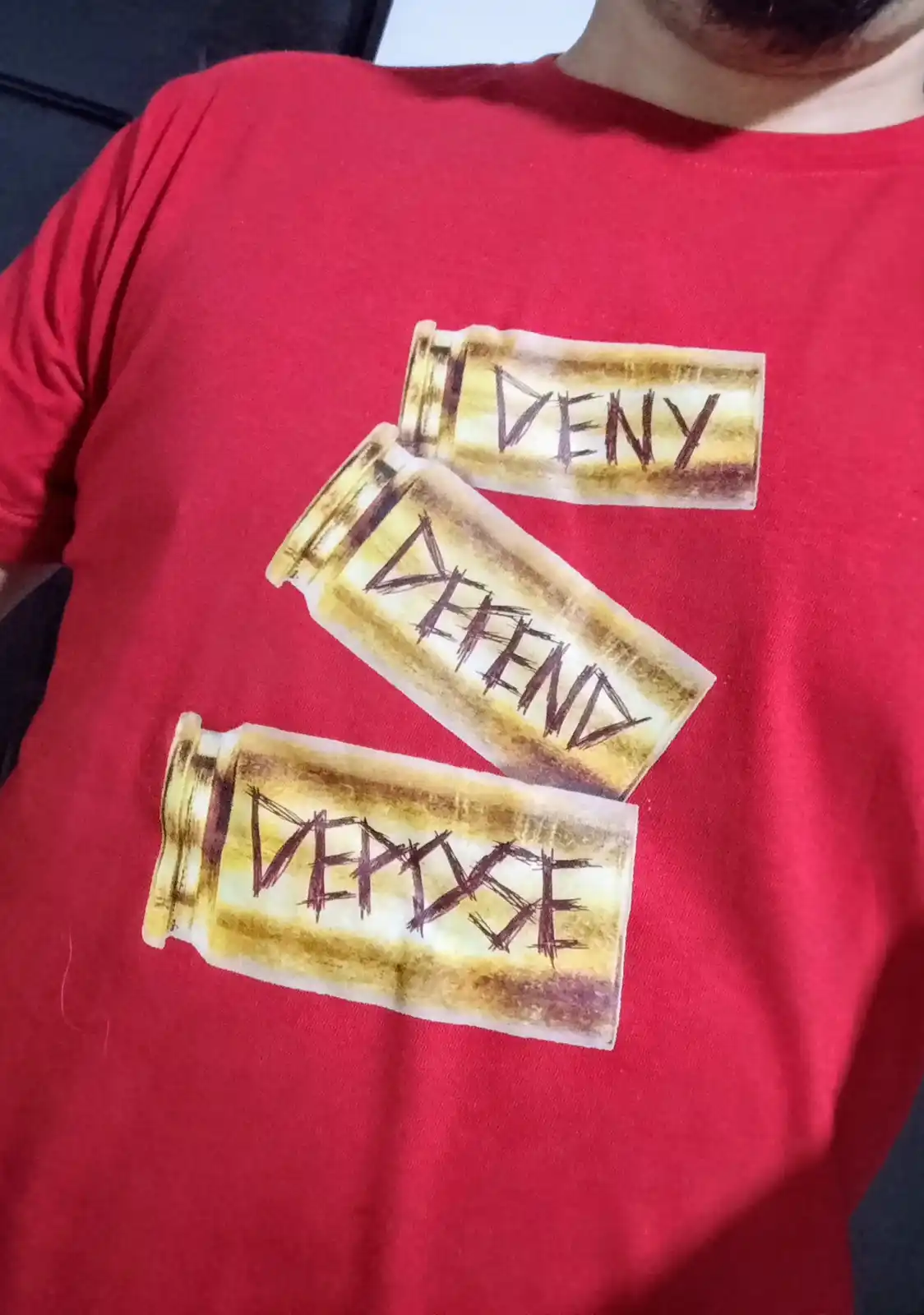 A person wearing a red t-shirt with three graphic bullet casings printed on the chest, each labeled with the words 'DENY'…