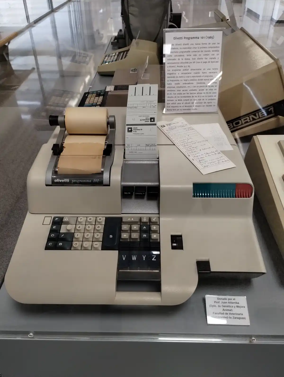 A beige Olivetti Programma 101 (1965) desktop computer is displayed in a glass case, featuring a keyboard, a paper roll…