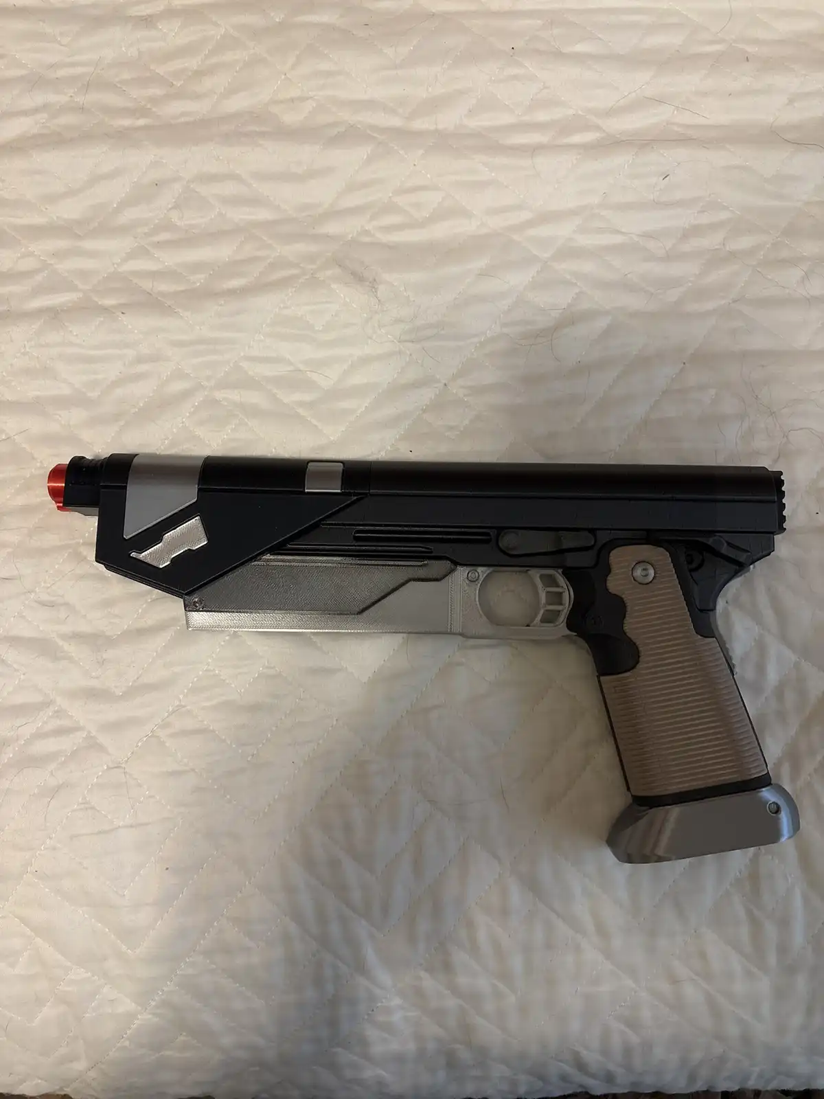 A 3d-printed replica of a futuristic pistol, featuring black, silver, and tan-colored parts with a red tip, lies…