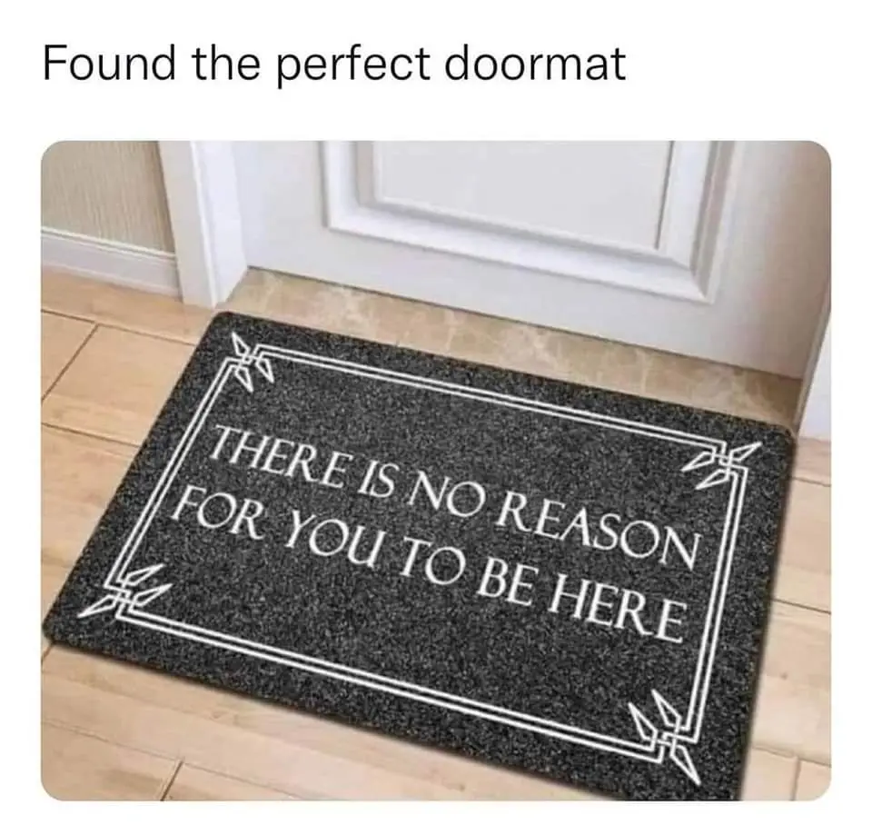 A black doormat with white text and decorative border sits on a wooden floor in front of a white door. The mat reads 'THERE…