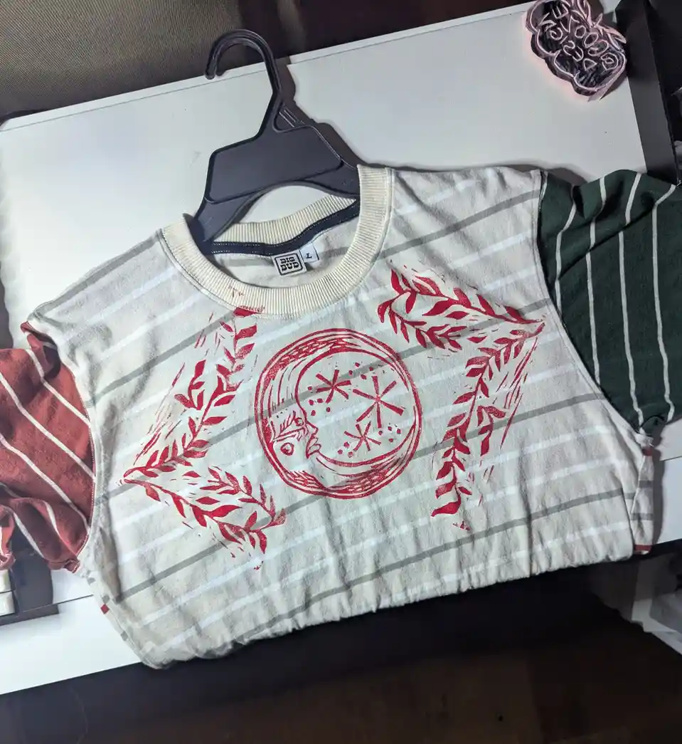 A striped, beige-and-gray sweater with a red printed design of a laurel wreath surrounding a circular motif with star-like…