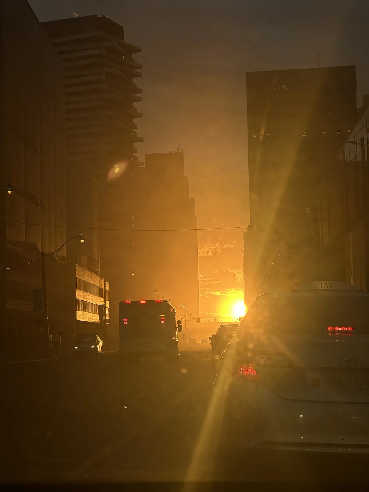 A hazy urban street scene at sunset, viewed from inside a car, with a taxi and bus ahead, the sun glowing orange between…