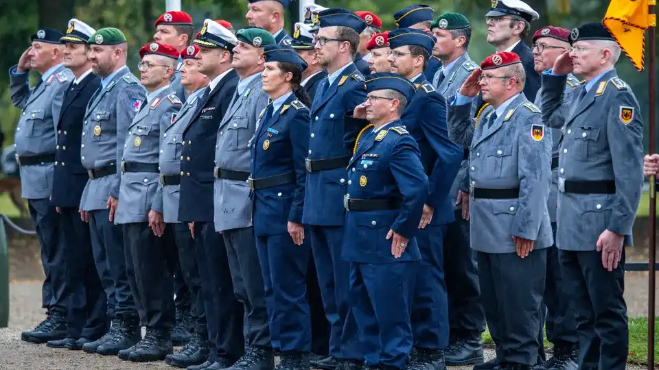 A line of German military personnel in uniform, mostly saluting, with one woman in the center not saluting and looking…