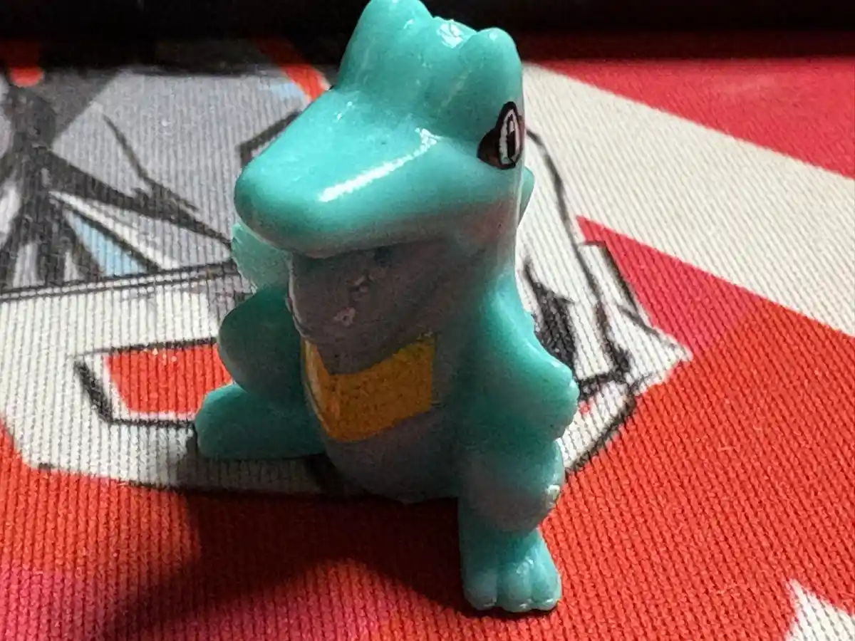 A small, teal-colored plastic figurine of the Pokémon Squirtle stands on a red and white fabric with a printed anime-style…