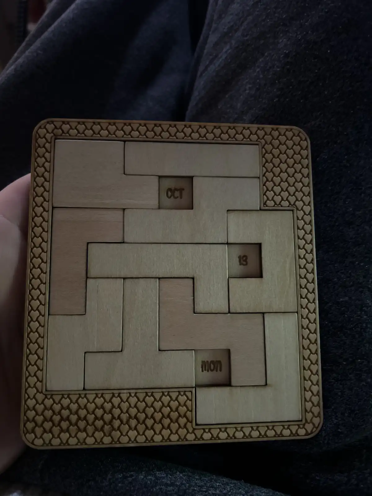 A hand holds a wooden puzzle calendar with interlocking pieces forming a grid; three small squares are visible with engraved…
