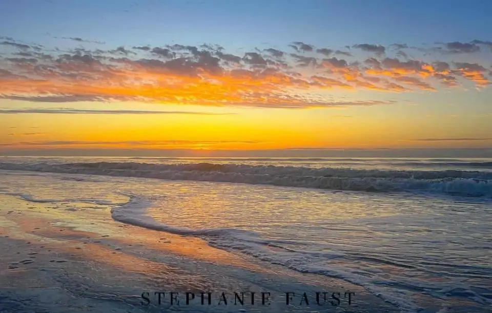 A serene beach sunset photo with orange and yellow clouds reflecting on the water, waves rolling in, and the photographer's…