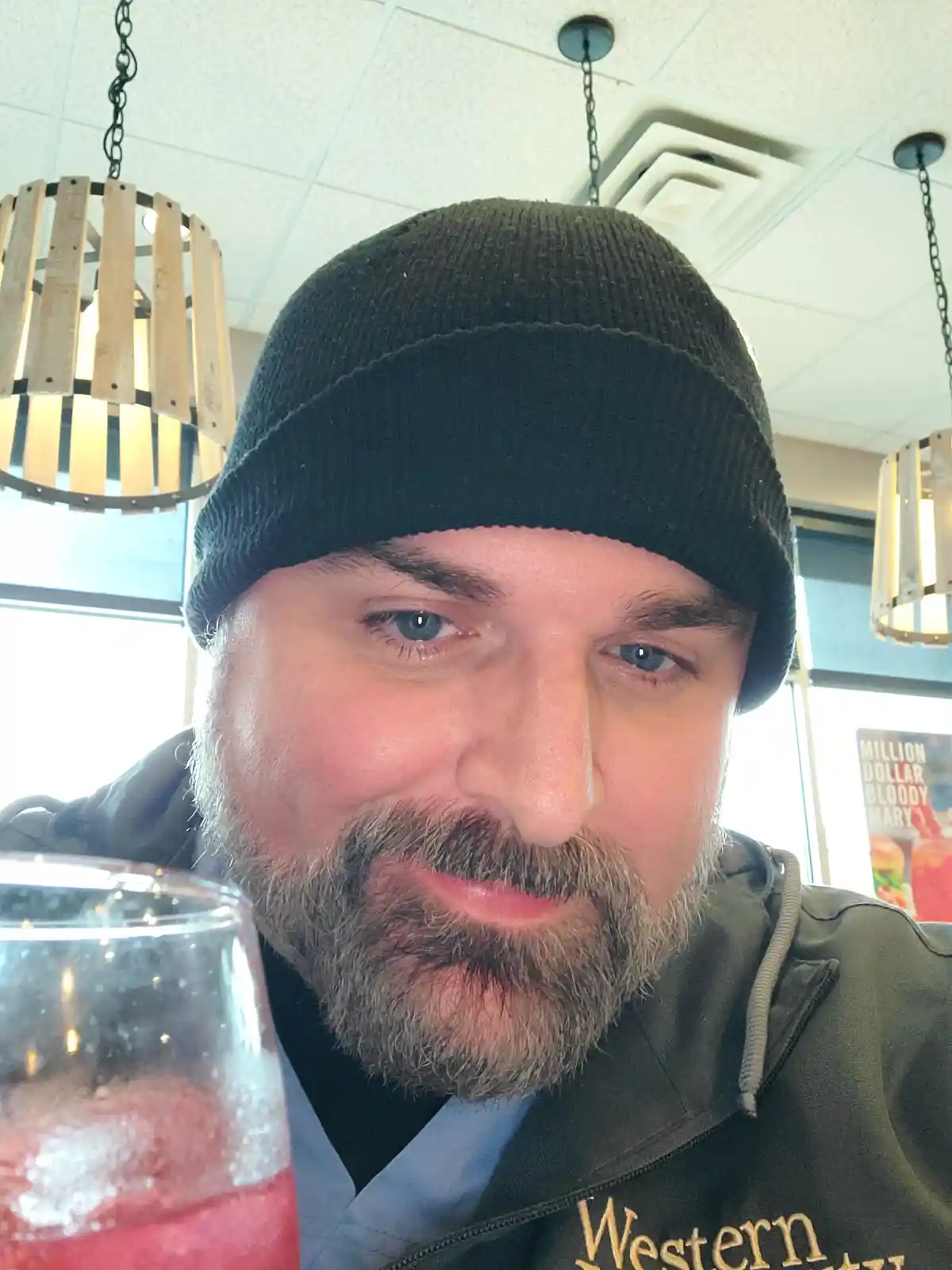 A man with a beard and black beanie stares intently at the camera while holding a glass of red drink with ice, likely a…