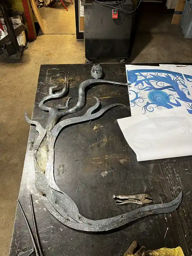 A metal sculpture of a stylized tree with twisted branches sits on a workshop table next to a blue and white paper design…