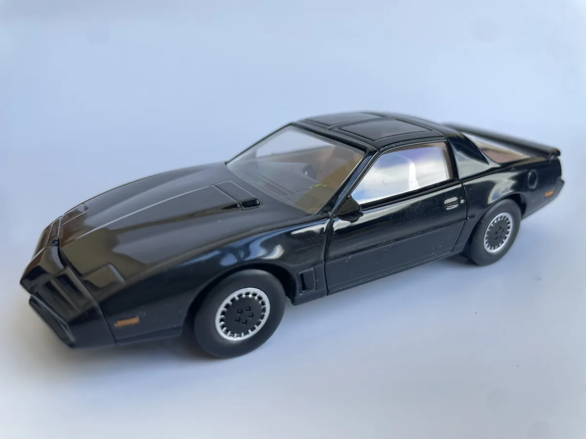 A black toy car, resembling the Pontiac Trans Am from Knight Rider, is photographed against a plain white background. The…