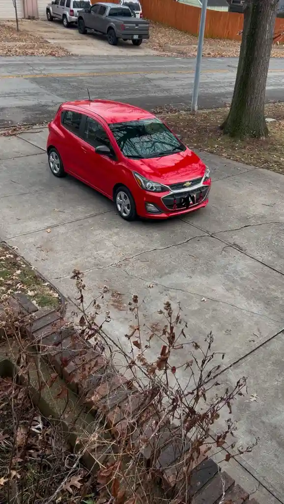 A bright red Chevrolet Spark is parked on a cracked concrete driveway, with a brick border and dry weeds in the foreground…
