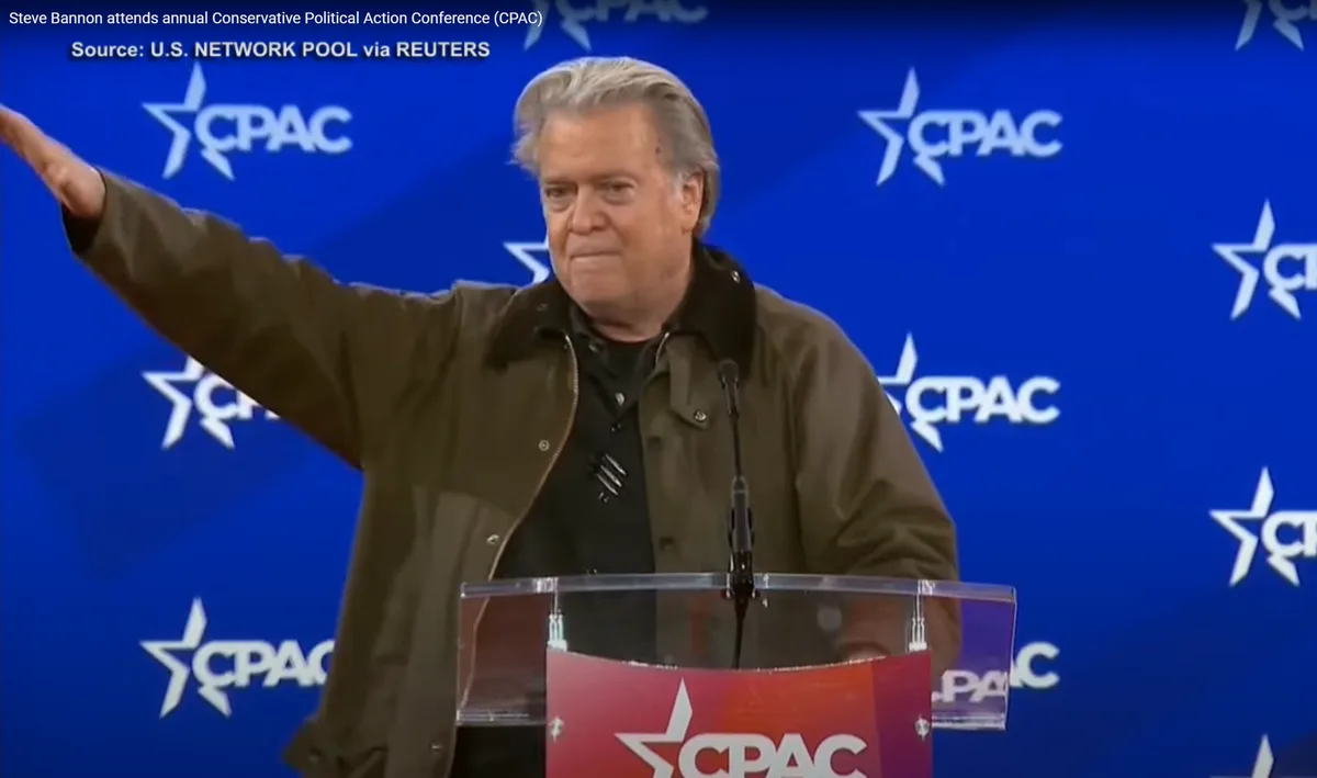 A man with gray hair, wearing a brown jacket over a black shirt, stands at a clear podium with a red CPAC logo, gesturing…