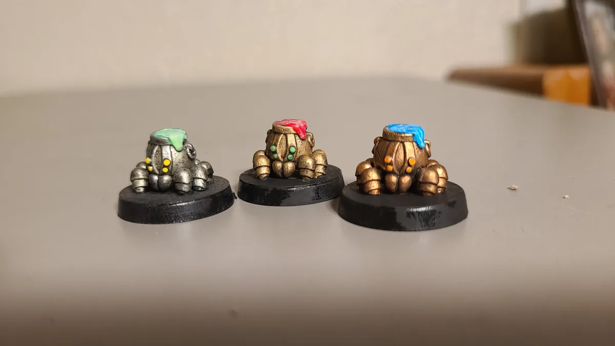 Three painted tabletop miniatures, each with a different colored paint blob (green, red, blue) dripping from their tops…