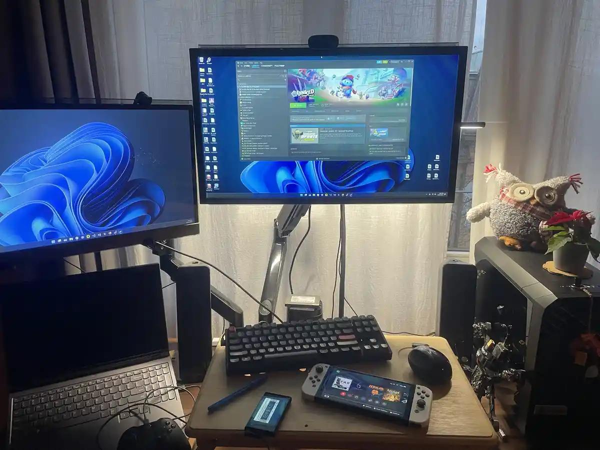A desk setup with two monitors, a laptop, a gaming console, and a PC tower. The top monitor displays the Steam library with…
