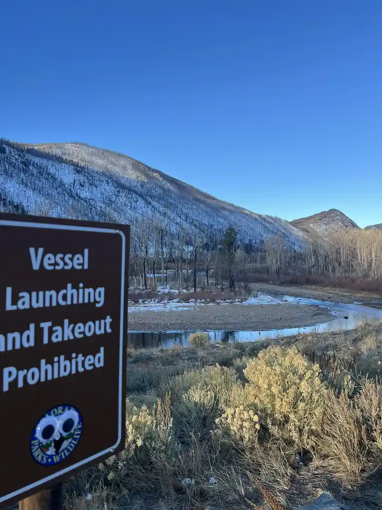 A brown sign with white text reading 'Vessel Launching and Takeout Prohibited' stands in front of a snowy mountain landscape…