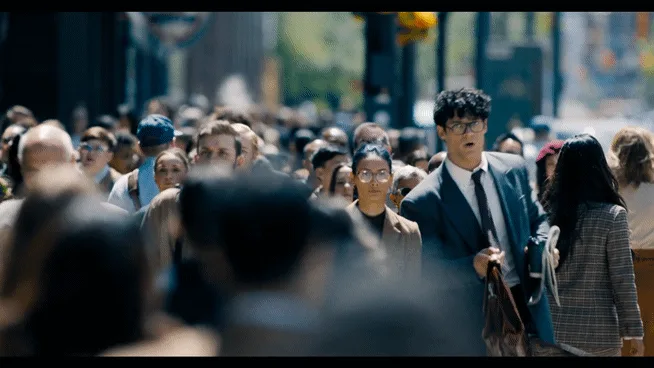 A man in a suit and glasses walks through a crowded city street, initially looking straight ahead with a serious expression…