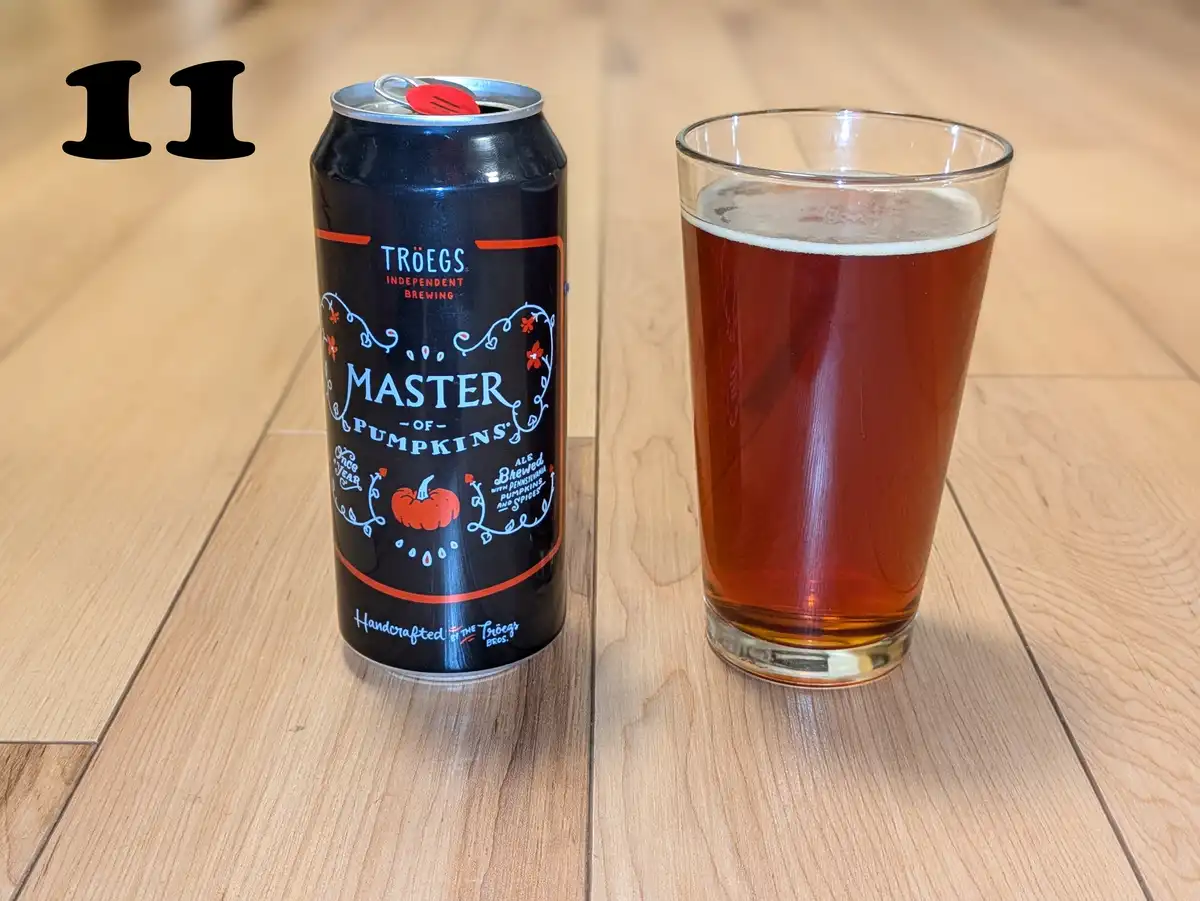 A black can of Troegs 'Master of Pumpkins' pumpkin ale sits next to a glass filled with the amber-colored beer on a light…