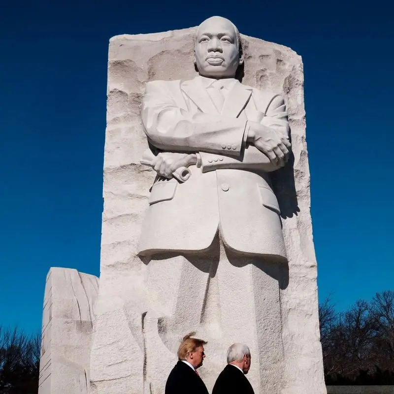 A large stone statue of Martin Luther King Jr. with arms crossed stands against a clear blue sky. At the base, two men, one…