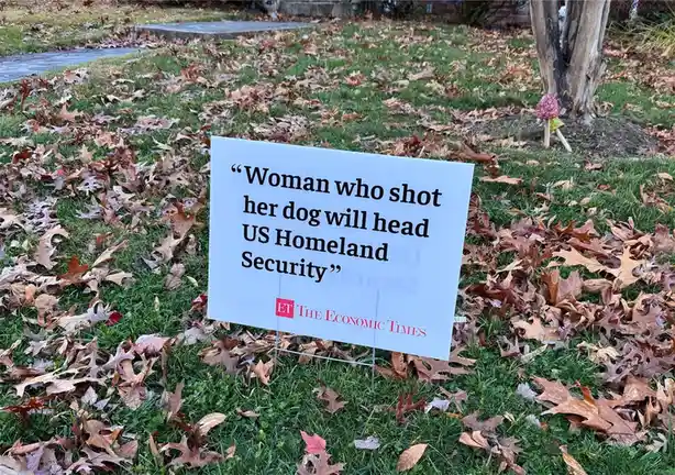 A white yard sign with black text reading 'Woman who shot her dog will head US Homeland Security' and a red 'ET THE ECONOMIC…