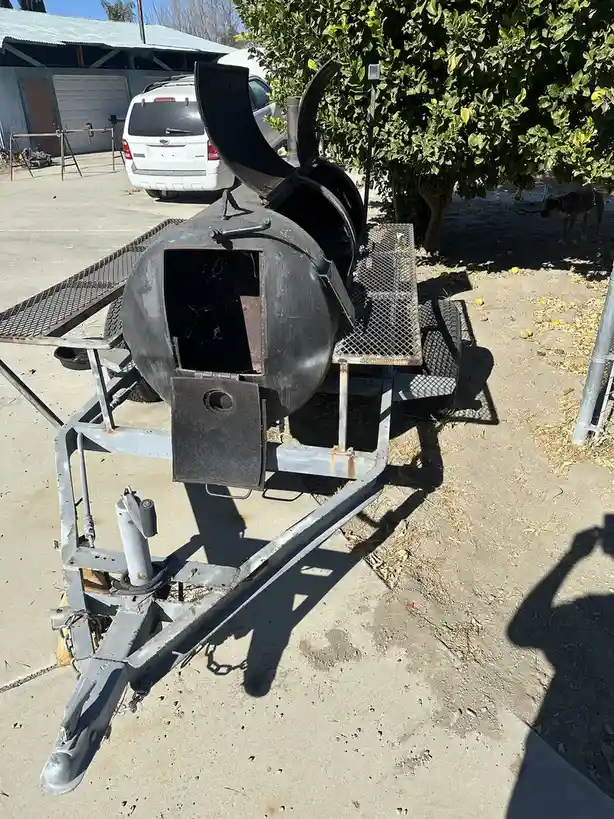 A large, black, barrel-shaped smoker on a metal trailer sits outdoors on a concrete driveway, its lid open and showing a…
