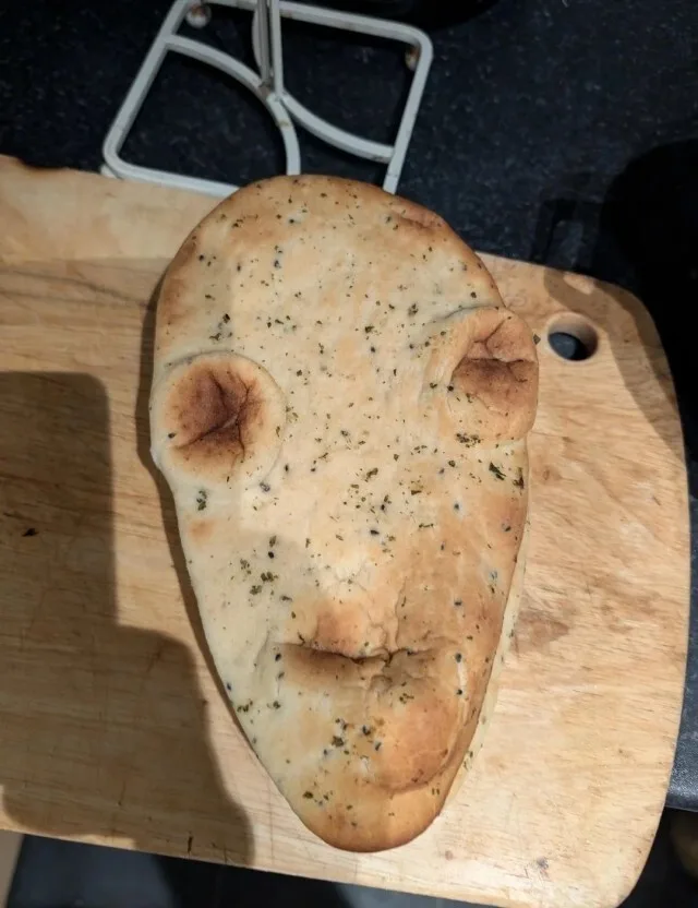 A piece of flatbread, seasoned with herbs and spices, rests on a wooden cutting board. Its shape and surface imperfections…