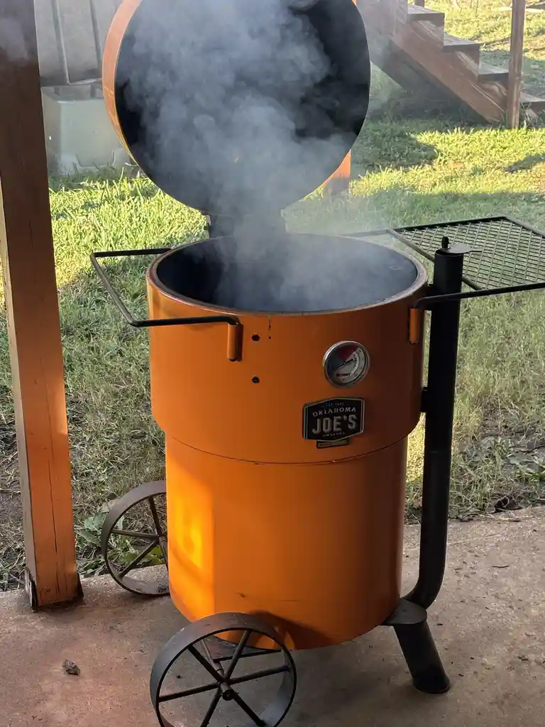 A large orange Oklahoma Joe's smoker with its lid open, emitting thick smoke, sits on a patio with grass and wooden stairs…