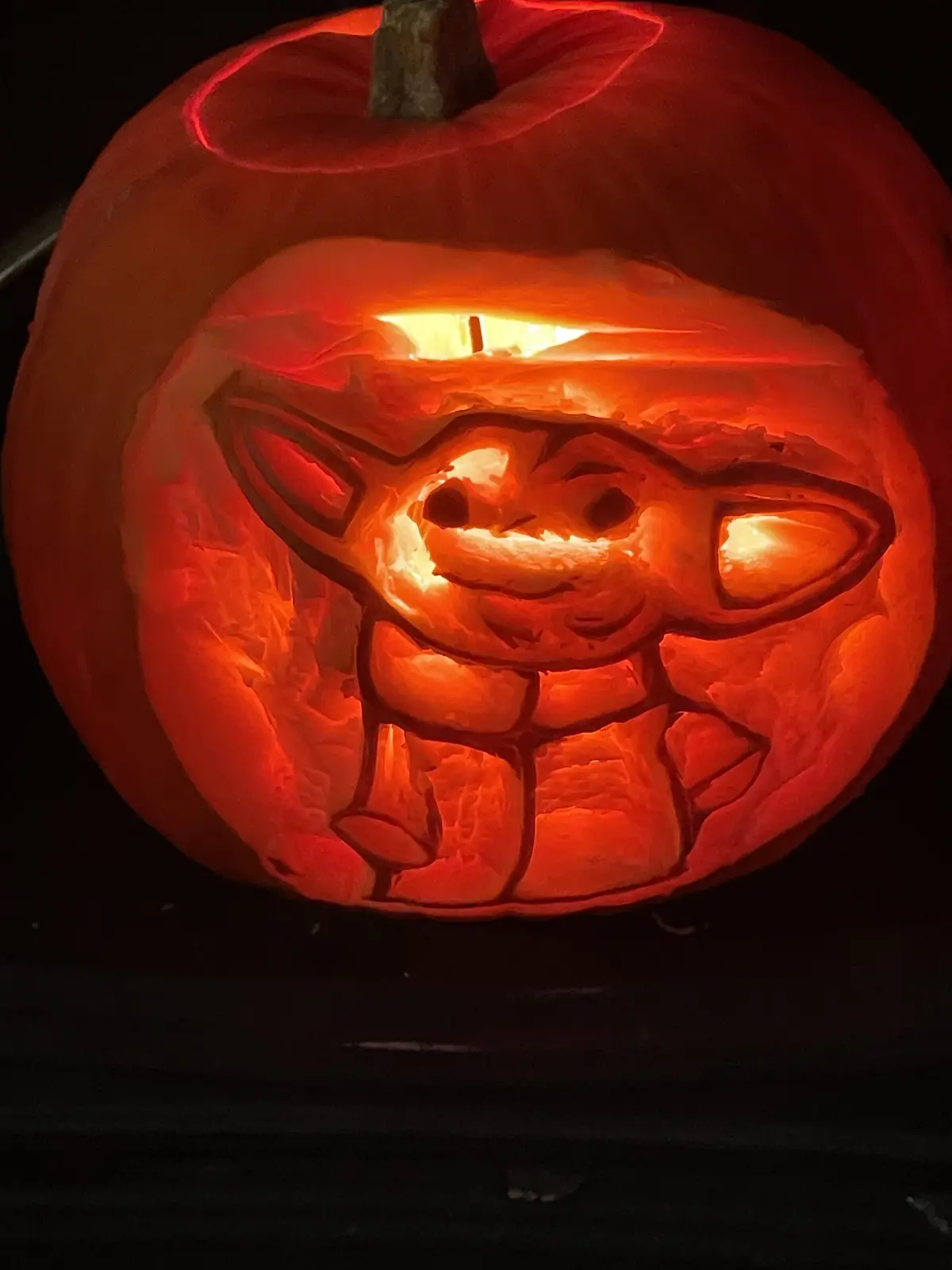 A close-up of a carved Halloween pumpkin illuminated from within, featuring a detailed design of the character Baby Yoda…