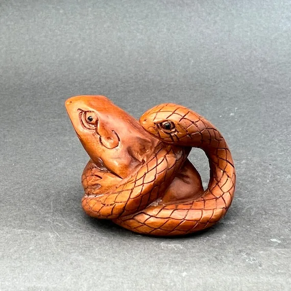 A wooden carving depicts a frog and a snake intertwined, with the snake coiled around the frog's body. The carving is…