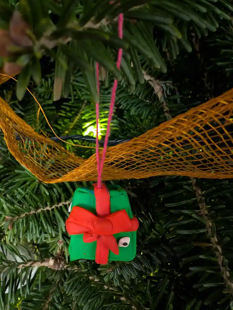 A handmade clay Christmas ornament shaped like a green gift box with a red bow and a single googly eye, hanging on a pine…