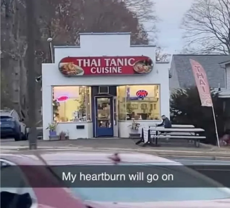 An exterior shot of a restaurant with a red sign reading 'THAI TANIC CUISINE', a play on 'Thai' and 'Titanic'. A person sits…