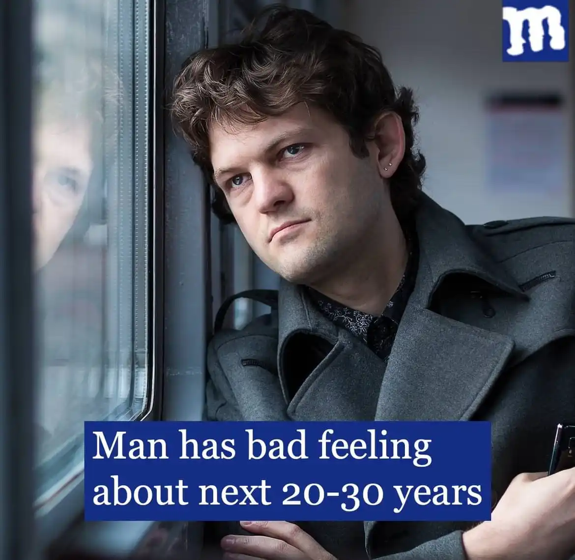 A man with a serious, worried expression leans against a train window, his reflection visible in the glass. He wears a grey…
