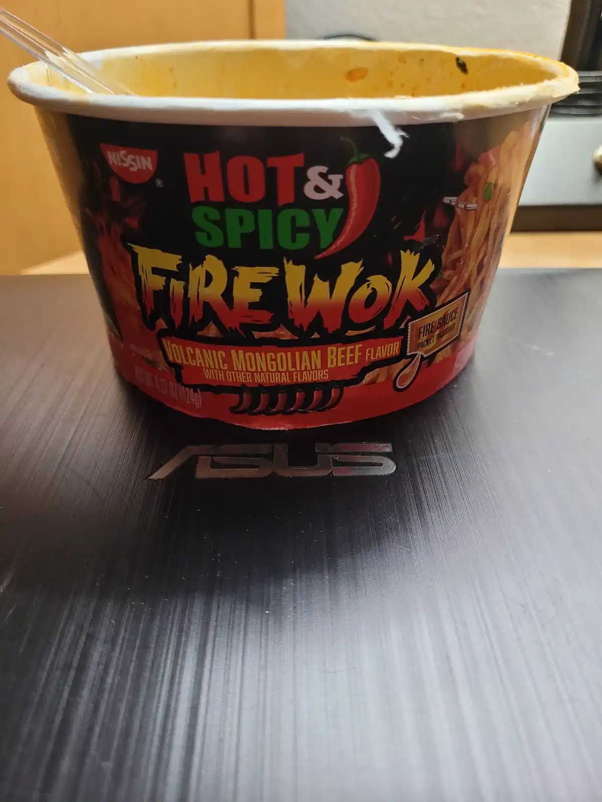 An empty Nissin Hot & Spicy Fire Wok cup, labeled 'Volcanic Mongolian Beef Flavor,' sits on a black ASUS laptop lid with the…