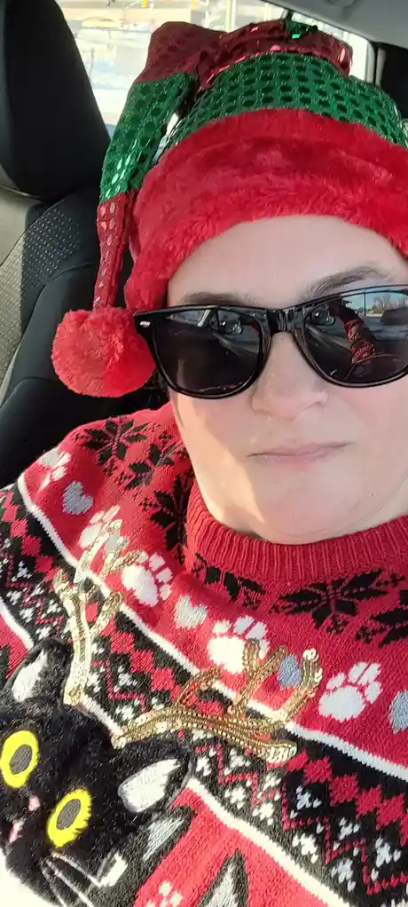 A person sits in a car wearing a red and green sparkly elf hat with a red pom-pom, black sunglasses, and a red, black, and…