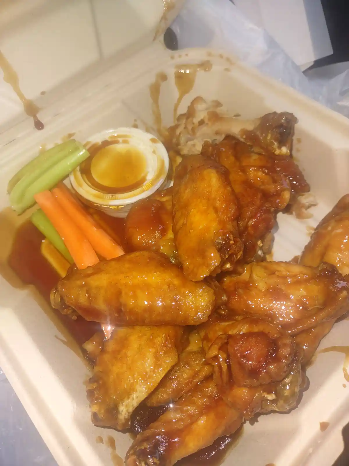 A close-up photo shows a white takeout container filled with glossy, sauce-covered chicken wings, some bones visible…