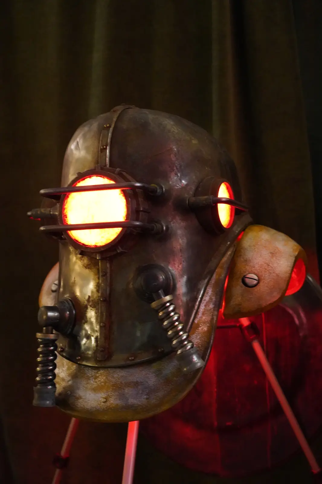 A close-up of a rusty, steampunk-style metal helmet with glowing red-orange lights for eyes and mouthpiece, mounted on a…