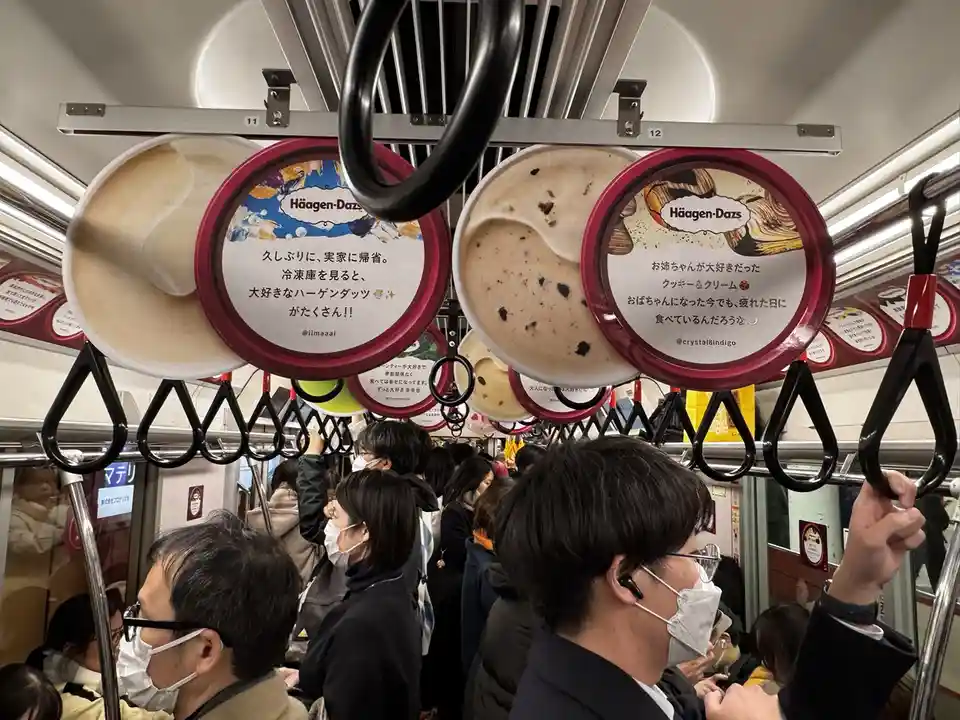 Inside a crowded Japanese train, people hold onto overhead handrails while large, circular Häagen-Dazs ice cream tubs hang…