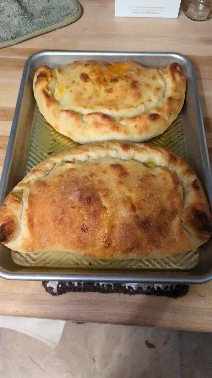 Two golden-brown, baked calzones sit on a metal baking sheet, resting on a wooden countertop. The calzones have puffy…